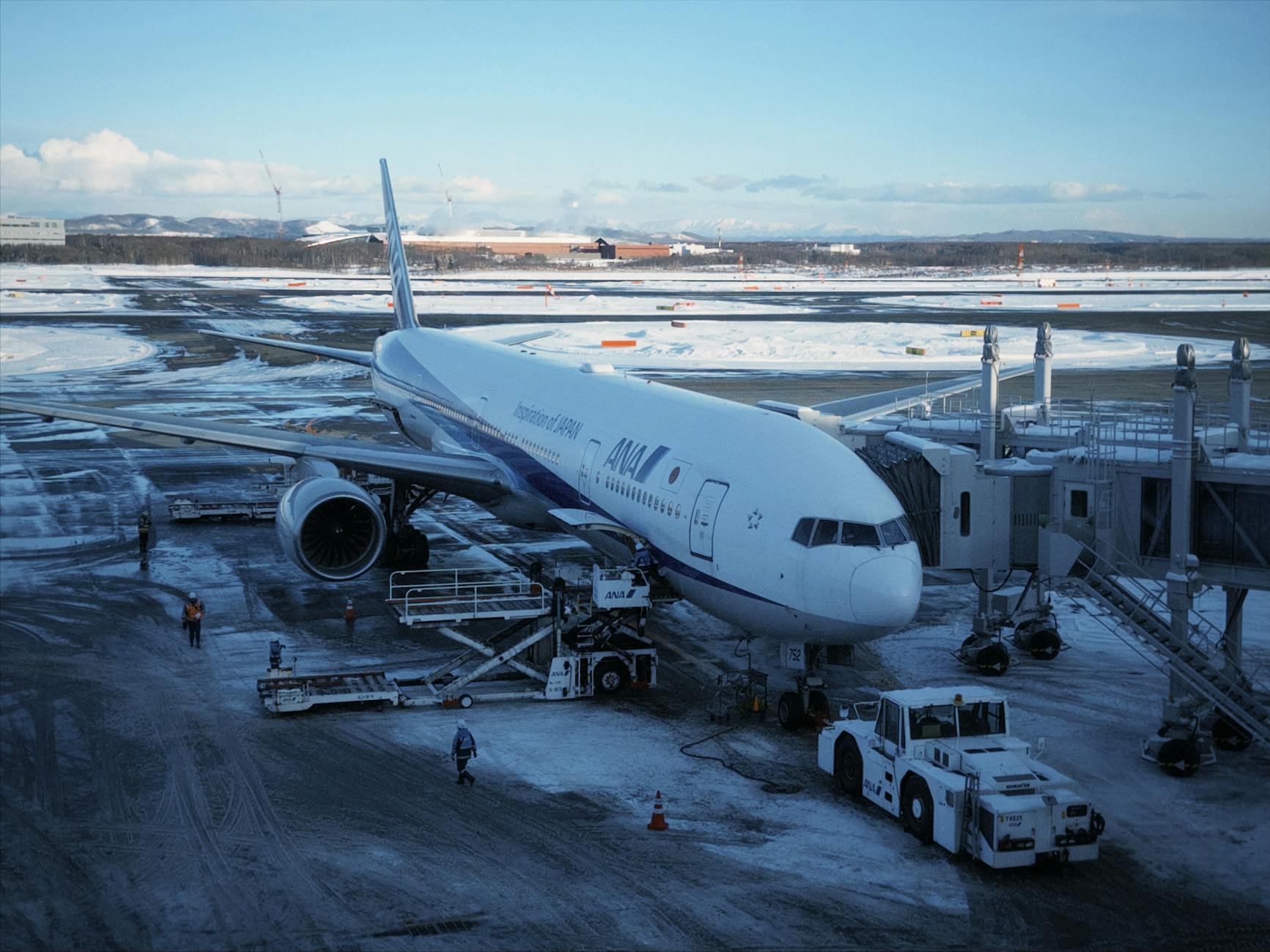An airplane parked at New Chitose Airport in winter, surrounded by snow and ground crew.